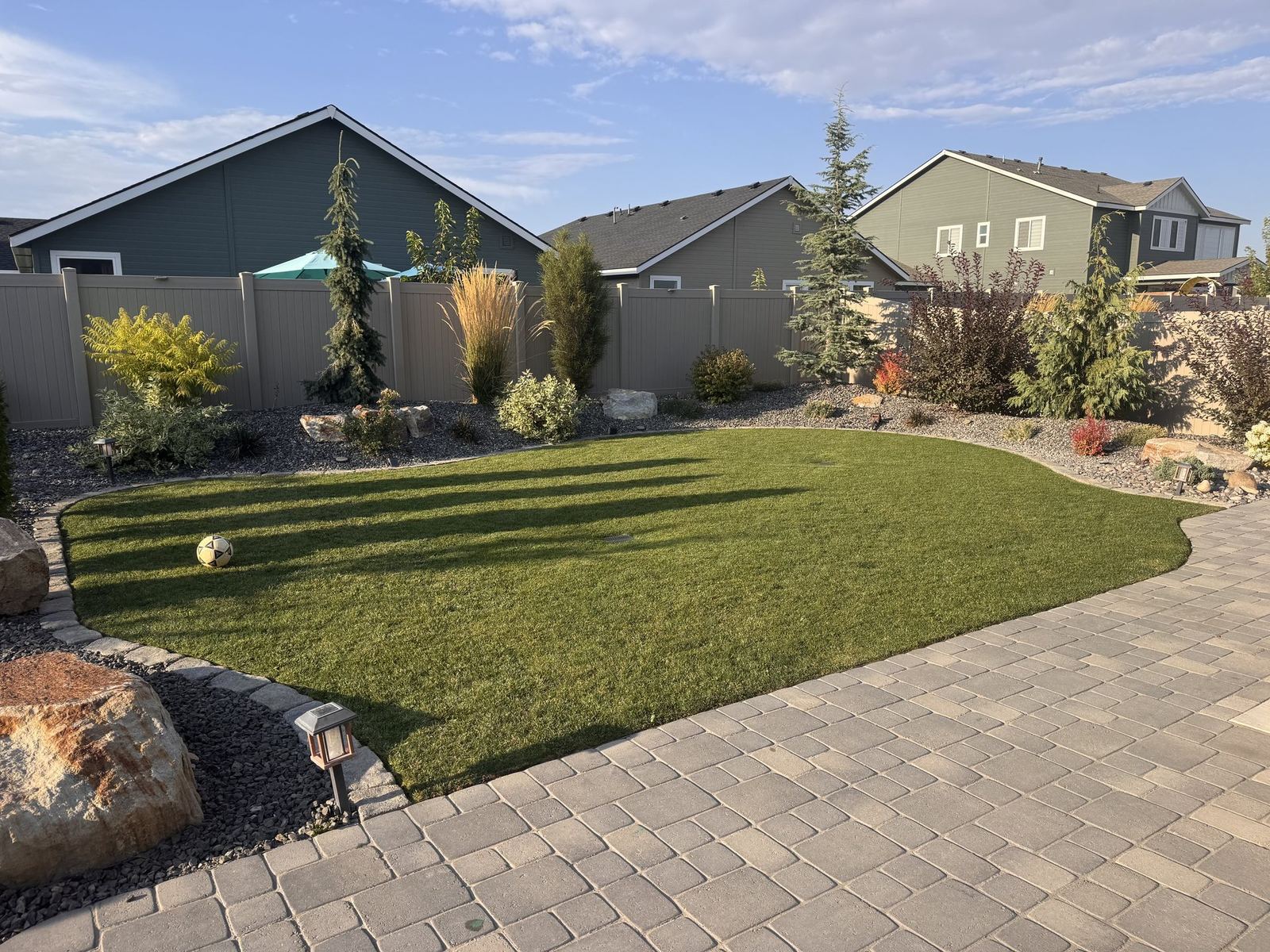 Finished backyard build by Cato Industries featuring a paver patio, artificial turf lawn, and perimeter rock landscaping at golden hour