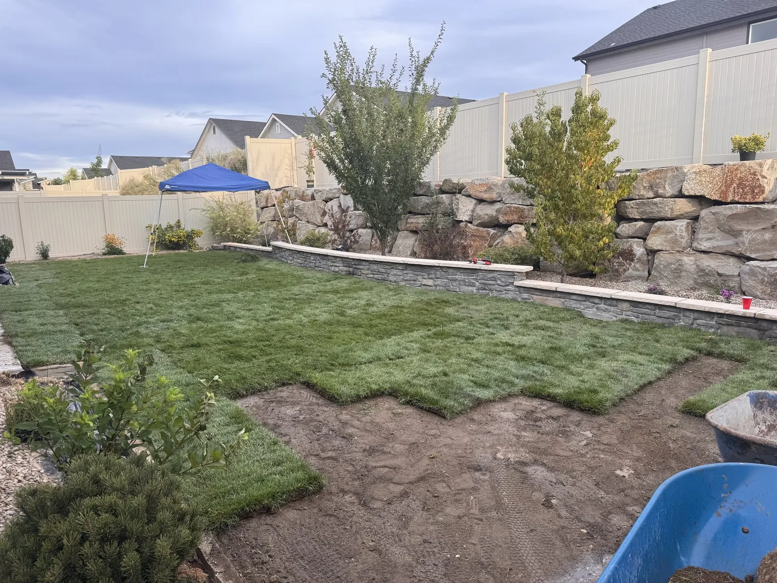 Backyard mid-build: boulder retaining wall in place, sod being laid, construction tools and canopy still on site